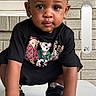 toddler, child, portrait, crouching, sitting, looking_at_camera, serious, black_shirt, bear_graphic, shoes, table, outdoor, brick_wall, window, house_exterior, smartphone_screenshot, phone_icons, vertical_photo, daylight, face
