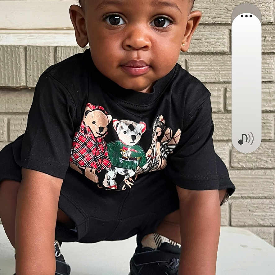 Cartrail is registered to the contest to win money with this photo: toddler, child, portrait, crouching, sitting, looking_at_camera, serious, black_shirt, bear_graphic, shoes, table, outdoor, brick_wall, window, house_exterior, smartphone_screenshot, phone_icons, vertical_photo, daylight, face