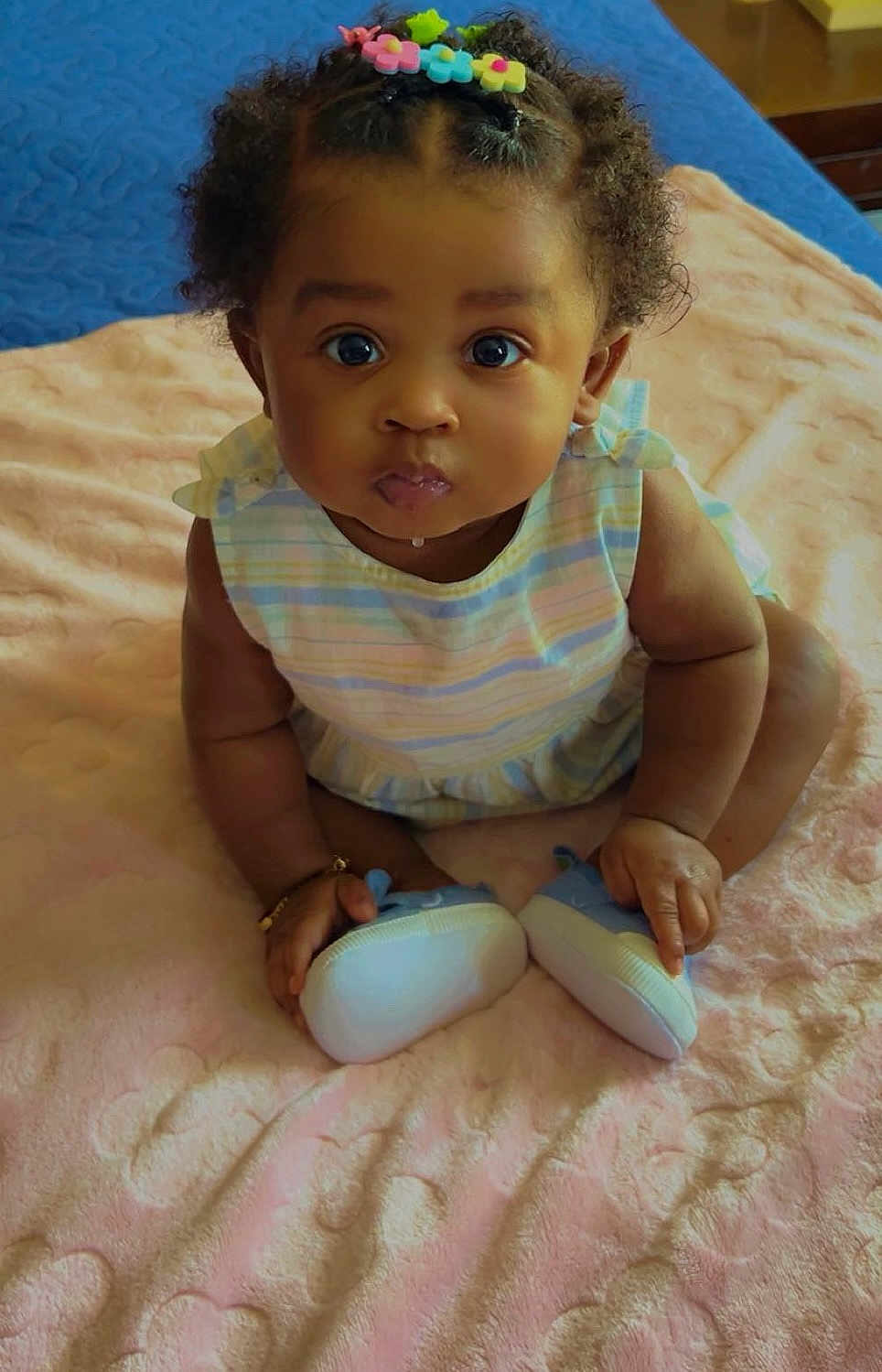 Nyla joined the competition — help win amazing prizes! baby, child, infant, cute, sitting, blue_shoes, hair_clips, curly_hair, pink_blanket, striped_dress, indoors, portrait, adorable, small_hands, bracelet, looking_up, soft_texture, closeup, young, innocent