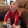 baby, child, plaid_shirt, jeans, couch, brown, pacifier, toy, flowers, vase, indoor, curious, sitting, looking_up, home, furniture, casual_clothing, hands, face, expression