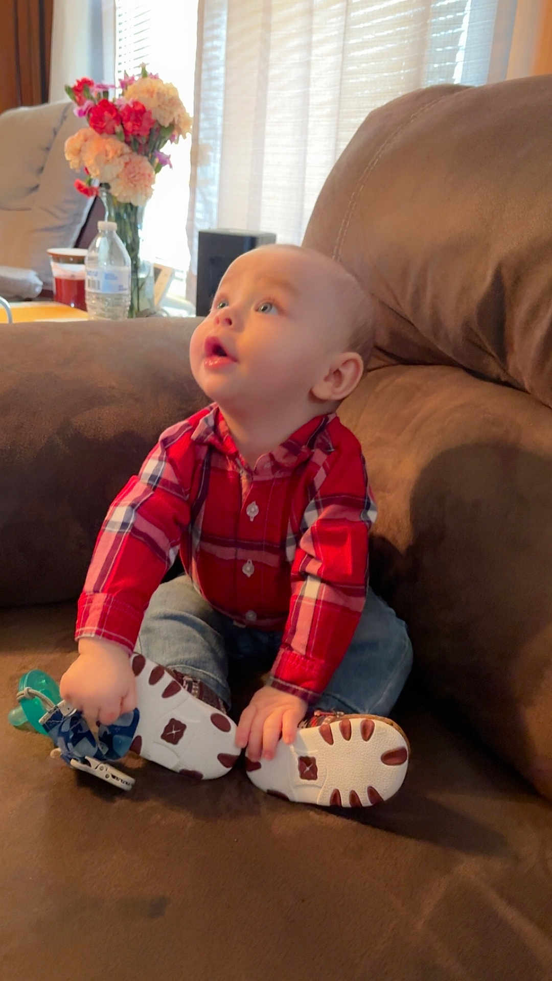Zechariah joined the competition — help win amazing prizes! baby, child, plaid_shirt, jeans, couch, brown, pacifier, toy, flowers, vase, indoor, curious, sitting, looking_up, home, furniture, casual_clothing, hands, face, expression