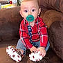baby, pacifier, red_shirt, plaid, blue_jeans, sitting, couch, brown_furniture, indoor, flowers, vase, water_bottle, child, cute, footwear, shoes, home, portrait, infant, cozy