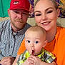 Zechariah is registered to the contest to win money with this photo: baby, spoon, man, woman, family, smile, indoor, plaid_shirt, red_cap, orange_shirt, curious, child, holding, portrait, happy, people, casual, background, wood, colorful