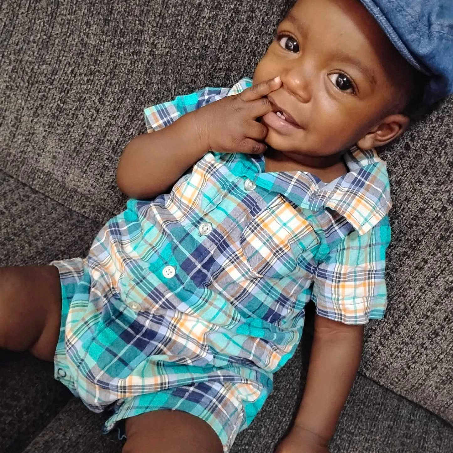 Kashton is registered to the contest to win money with this photo: baby, blue_cap, casual_outfit, child, comfortable, couch, curious_eyes, cute, fabric_texture, hand_near_face, indoor, plaid_clothing, playful, portrait, relaxed, seated, skin, smiling, toddler, young_child