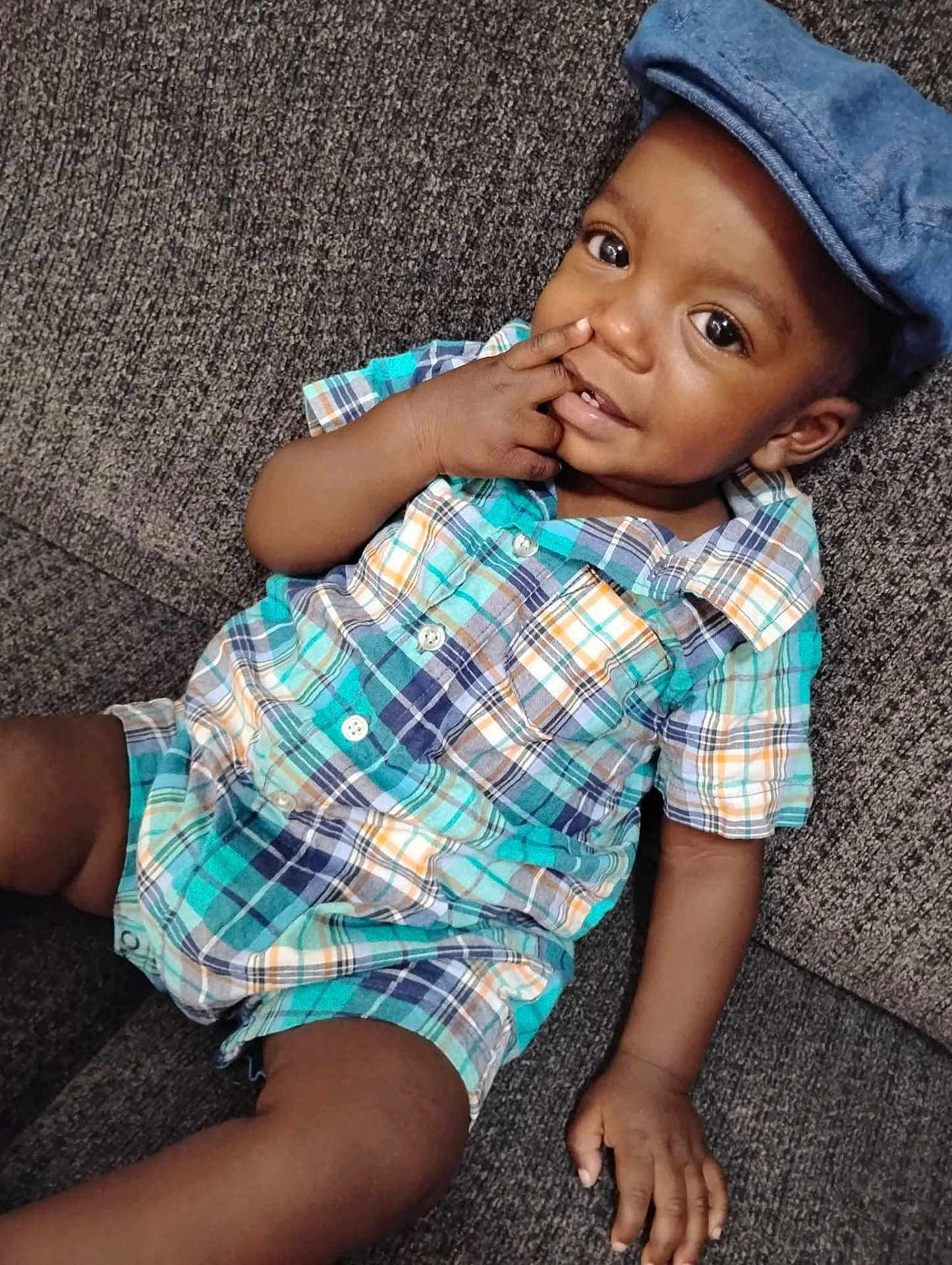 Kashton is registered to the contest to win money with this photo: toddler, child, plaid_clothing, blue_cap, couch, indoor, portrait, smiling, curious_eyes, hand_near_face, casual_outfit, baby, seated, cute, relaxed, fabric_texture, skin, young_child, playful, comfortable