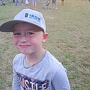 Hunter is registered to the contest to win money with this photo: boy, child, smiling, cap, hat, gray_clothing, chain_necklace, outdoor, grass, festival, crowd, people, fair, carnival, stroller, casual_wear, young, happy, portrait, daytime