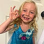 child, girl, blonde_hair, blue_dress, face_paint, hand_gesture, smiling, teeth, door, doorknob, indoor, portrait, happy, cute, young, long_hair, sparkle, hand, gesture, seahorse_tattoo