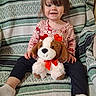 toddler, child, smiling, stuffed_animal, plush_toy, dog_toy, red_bow, heart_decorations, patterned_shirt, dark_pants, white_socks, blanket, green, white, striped_blanket, indoor, seated, happy, cute, portrait