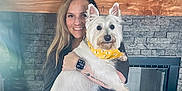Berkley The Barkster joined the competition — help win amazing prizes! woman, dog, indoor, fireplace, stone_wall, wooden_mantel, yellow_bandana, smiling, holding, pet, white_dog, tattoo, long_hair, watch, plant, framed_photo, home_decor, casual_clothing, cozy, portrait