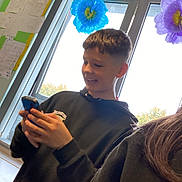 Crackwhore is registered to the contest to win money with this photo: boy, child, teenager, smiling, smartphone, hoodie, school_uniform, badge, hands, short_hair, window, paper_flowers, classroom, crafts, poster, indoor, daylight, desk, glass_window, casual