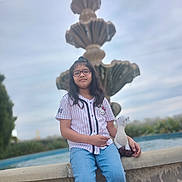 Delilah is registered to the contest to win money with this photo: child, girl, glasses, plush_toy, fountain, water, outdoor, casual_clothing, jeans, short_sleeve_shirt, nature, sky, cloudy, portrait, sitting, relaxed, daytime, young, scenery, background_blur