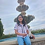 child, girl, glasses, plush_toy, fountain, water, outdoor, casual_clothing, jeans, short_sleeve_shirt, nature, sky, cloudy, portrait, sitting, relaxed, daytime, young, scenery, background_blur