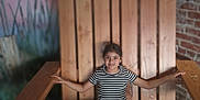 Delilah is registered to the contest to win money with this photo: child, striped_shirt, jeans, wooden_chair, oversized_chair, indoor, brick_wall, mural, bird, sunset, smiling, casual, relaxed, person, seated, portrait, playful, happy, feet_up, wooden_furniture