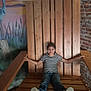 child, striped_shirt, jeans, wooden_chair, oversized_chair, indoor, brick_wall, mural, bird, sunset, smiling, casual, relaxed, person, seated, portrait, playful, happy, feet_up, wooden_furniture
