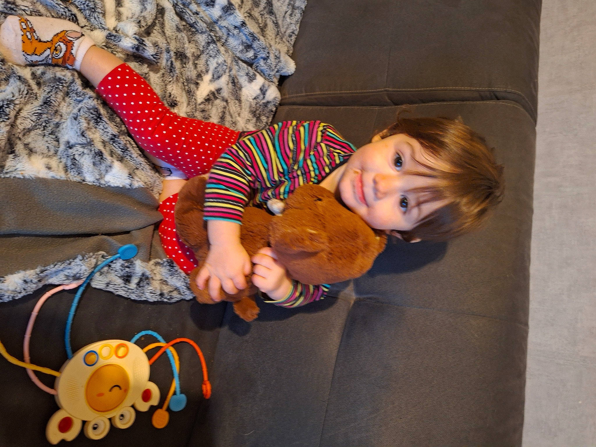 Lou participe au concours pour gagner de l'argent avec cette photo : baby_toddler_clothing, child, comfort, flooring, fun, happy, lap, linens, orange, pattern, person, room, sitting, smile, sock, stuffed_toy, textile, toddler, toy, tree