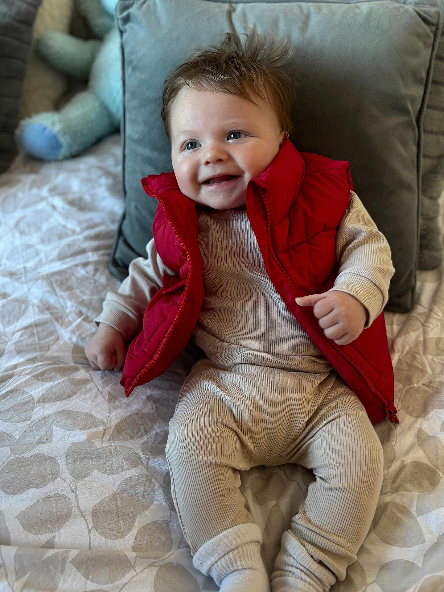 Albie is registered to the contest to win money with this photo: baby, child, smiling, red_vest, beige_outfit, bedspread, pillow, plush_toy, indoor, portrait, happy, cute, infant, sitting, cozy, clothing, face, person, messy_hair, soft_lighting