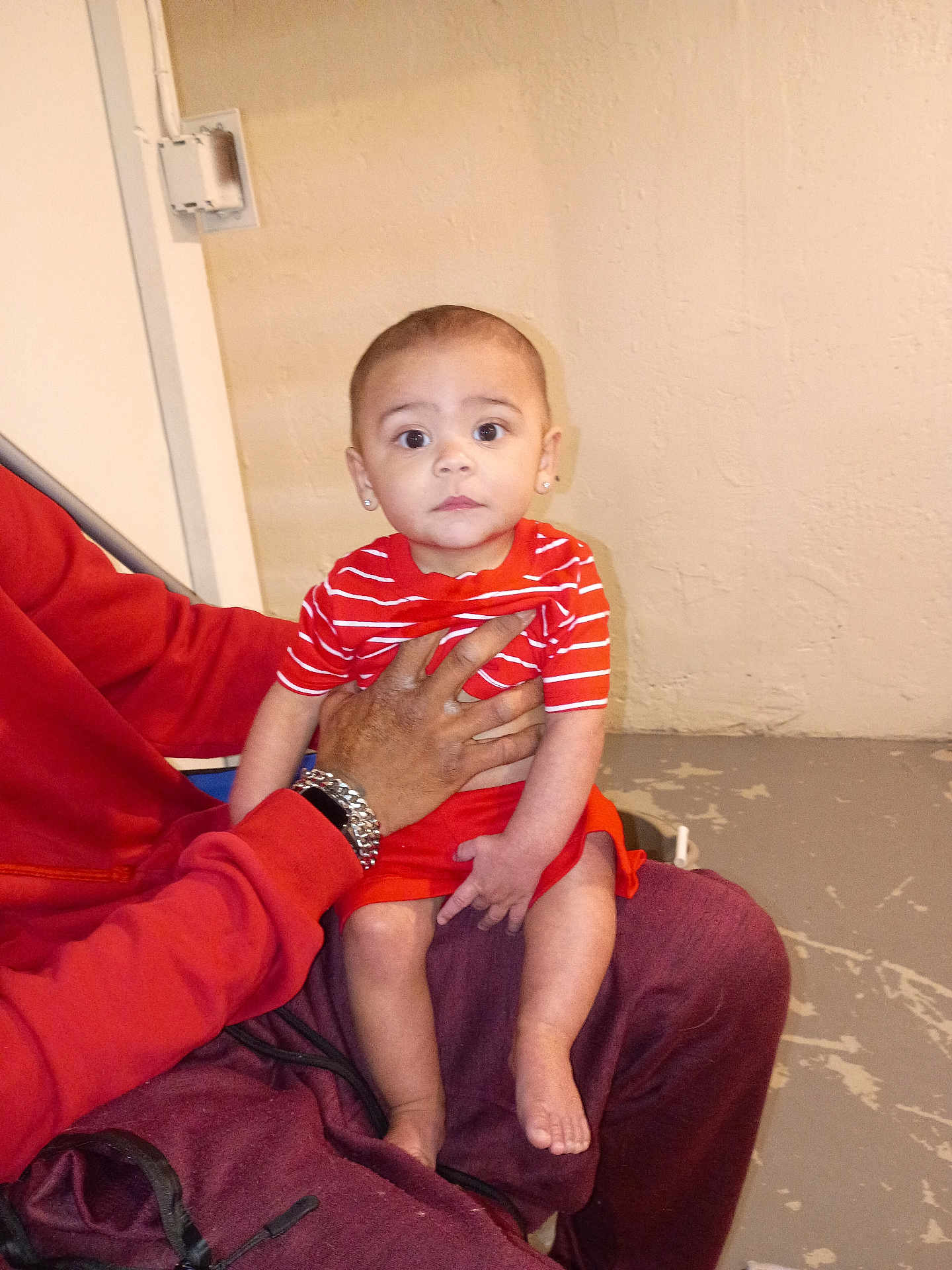 Lashawn joined the competition — help win amazing prizes! baby, child, person, striped_clothing, red_clothing, earrings, adult_hand, indoor, wall, floor, pants, jacket, sitting, holding, looking_at_camera, barefoot, expressionless, casual, portrait, human