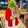 grinch_costume, baby, child, pajamas, fire_truck_pattern, person, restaurant, indoor, window, night, exit_sign, table, chair, booth_seating, floor_tile, wet_floor_sign, costume, holiday, green_face, red_clothing