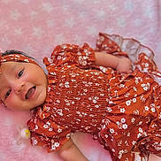 Dorothea is registered to the contest to win money with this photo: baby, infant, smiling, headband, floral_pattern, orange_clothing, pink_blanket, cute, happy, lying_down, soft_texture, child, portrait, indoors, apparel, headwear, skin, face, small_hand, pacifier