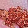 baby, infant, smiling, headband, floral_pattern, orange_clothing, pink_blanket, cute, happy, lying_down, soft_texture, child, portrait, indoors, apparel, headwear, skin, face, small_hand, pacifier