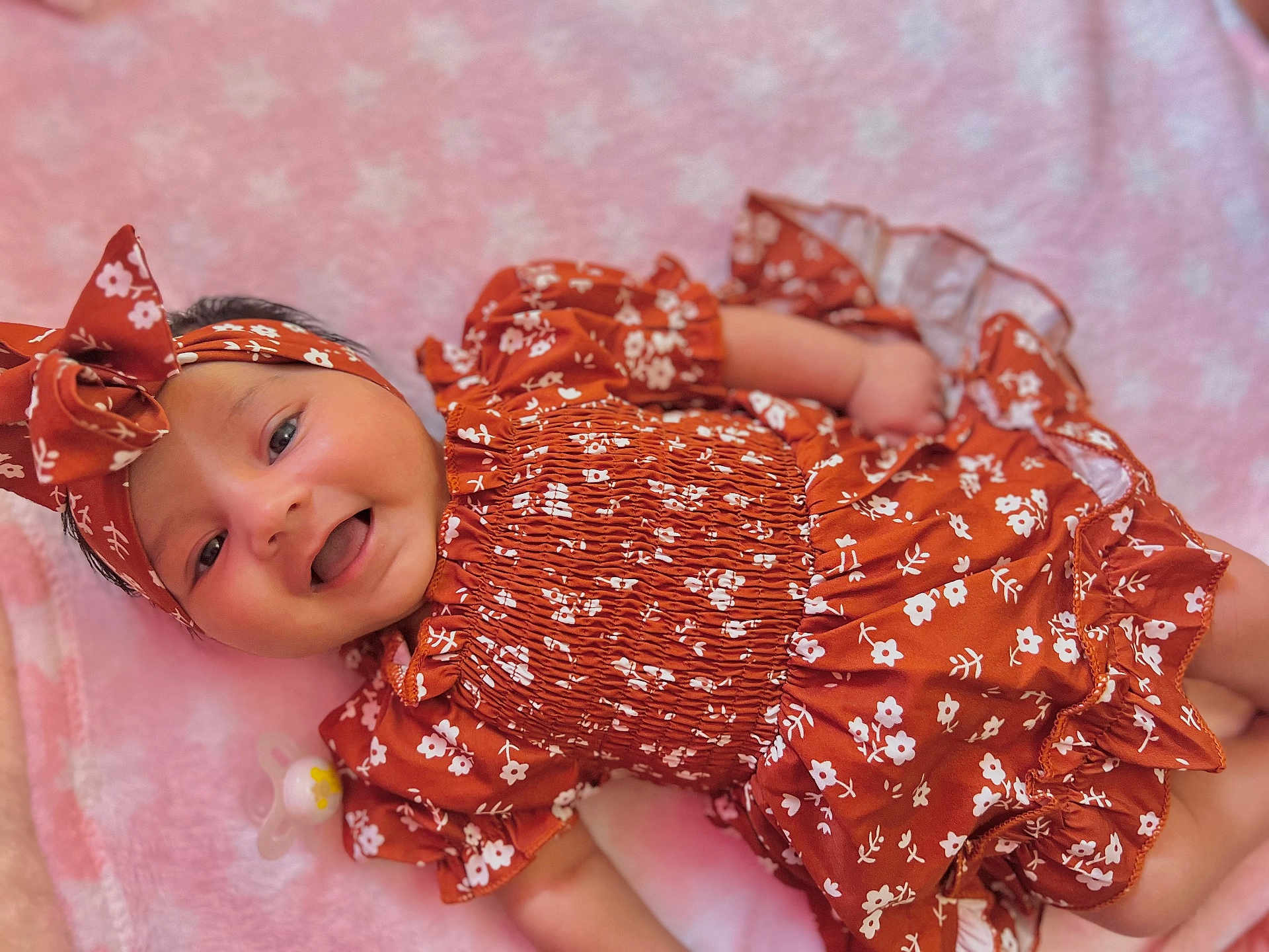 Dorothea is registered to the contest to win money with this photo: baby, infant, smiling, headband, floral_pattern, orange_clothing, pink_blanket, cute, happy, lying_down, soft_texture, child, portrait, indoors, apparel, headwear, skin, face, small_hand, pacifier