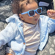 Eliana is registered to the contest to win money with this photo: baby, child, sunglasses, blue_jacket, curly_hair, cushion, patterned_cushion, sunny_day, outdoor, portrait, fashion, casual_clothing, light_blue, relaxed, cute, infant, person, headwear, smile, baby_clothes