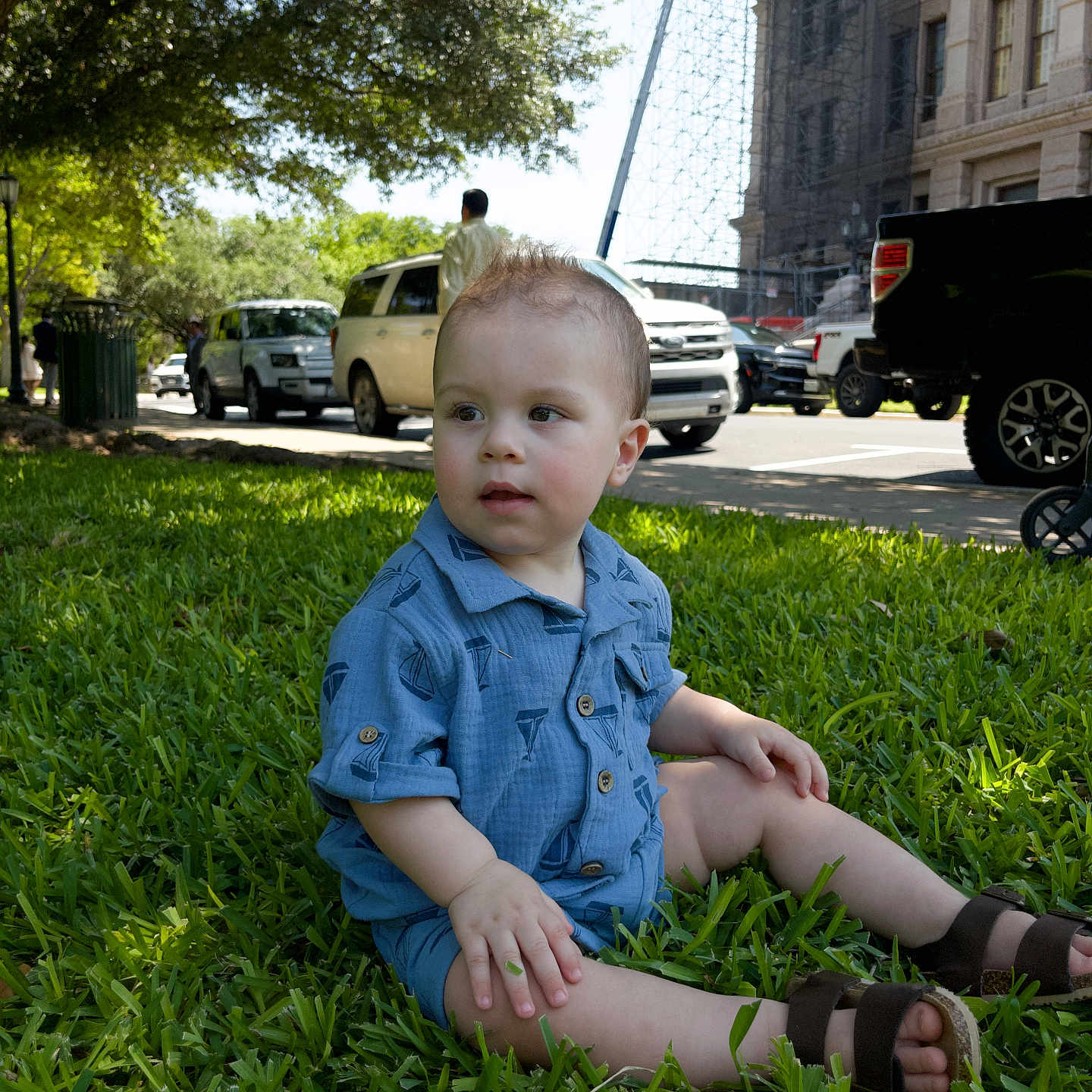 Ian is registered to the contest to win money with this photo: baby, blue_shirt, building, car, child, curious, daylight, grass, grass_field, outdoor, person, sandals, shoes, side_view, sitting, street, sunlight, toddler, tree, urban