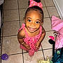 A'Myri is registered to the contest to win money with this photo: toddler, child, smiling, pink_dress, bow, curly_hair, floor_tiles, clothing, indoor, happy, baby, bracelet, playful, person, portrait, cute, young_child, looking_up, footwear, messy_room