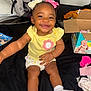 baby, child, smiling, sitting, yellow_shirt, white_socks, bows, hair_buns, diaper_box, car_seat, black_background, happy, indoor, cute, infant, clothing, person, toddler, toy, fabric