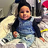 Malani is registered to the contest to win money with this photo: baby, child, denim_overalls, black_top, head_wrap, sitting, bed, blanket, car_seat, indoor, footwear, curious, cute, clothing, person, portrait, cozy, furniture, childrens_fashion, smiling