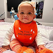 Malani joined the competition — help win amazing prizes! baby, child, bib, orange, headband, smile, striped_pants, slippers, bed, blanket, indoor, clothing, face, person, portrait, cute, halloween, cozy, toy, footwear