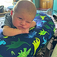 Koda is registered to the contest to win money with this photo: baby, infant, dinosaur_pillow, blanket, bedroom, window, wooden_door, striped_clothing, face, expression, indoor, resting, child, person, soft_light, cozy, toddler, skin, head, pose