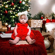 Echo is registered to the contest to win money with this photo: baby, child, red_dress, white_bow, headband, christmas_tree, ornaments, lights, holiday, festive, decorations, red_blanket, wooden_sled, indoors, cute, smiling, sitting, floor, cozy, celebration