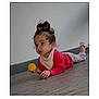 baby, child, floor, yellow_ball, red_shirt, white_pants, top_knot, indoor, curious, person, young_child, crawling, headwear, long_sleeves, footwear, wall, wooden_floor, play, infant, portrait