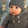baby, child, knit_hat, pom_poms, gray_sweater, blue_blanket, indoor, toy, curious, sitting, cute, infant, warm_clothing, face, eyes, buttoned_sweater, soft_toys, wooden_frame, cozy, portrait