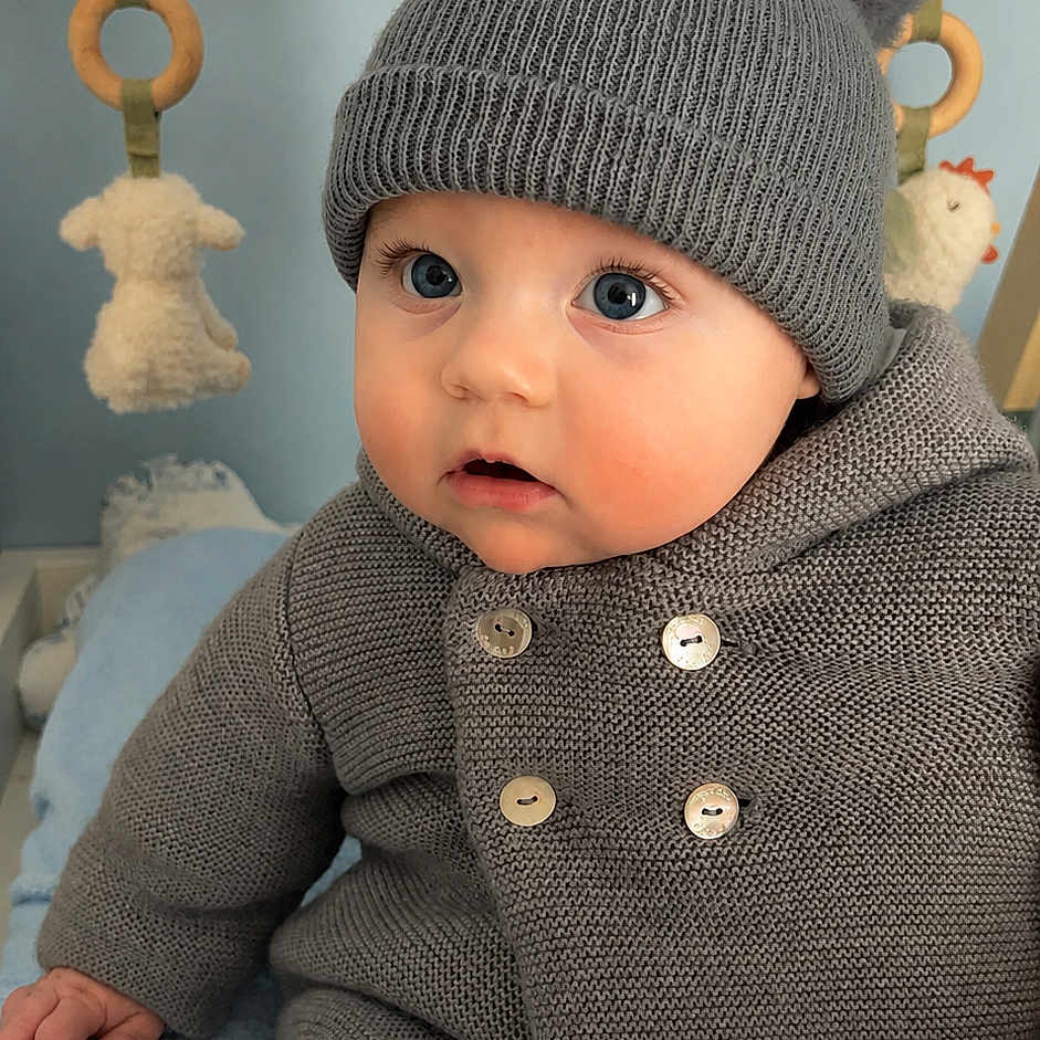 Raphaël participe au concours pour gagner de l'argent avec cette photo : baby, child, knit_hat, pom_poms, gray_sweater, blue_blanket, indoor, toy, curious, sitting, cute, infant, warm_clothing, face, eyes, buttoned_sweater, soft_toys, wooden_frame, cozy, portrait