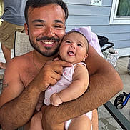Emersyn is registered to the contest to win money with this photo: man, baby, smile, outdoor, patio, chair, shirtless, beard, onesie, holding, happy, person, tattoo, window, sign, concrete_floor, arm, hand, family, baby_clothing
