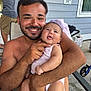 man, baby, smile, outdoor, patio, chair, shirtless, beard, onesie, holding, happy, person, tattoo, window, sign, concrete_floor, arm, hand, family, baby_clothing
