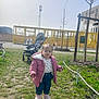 child, toddler, pigtails, pink_jacket, pink_boots, jeans, stroller, playground, slide, grass, park, rope_fence, tree, building, sunny_day, shadow, face, walking, winter_coat, outdoor_fun