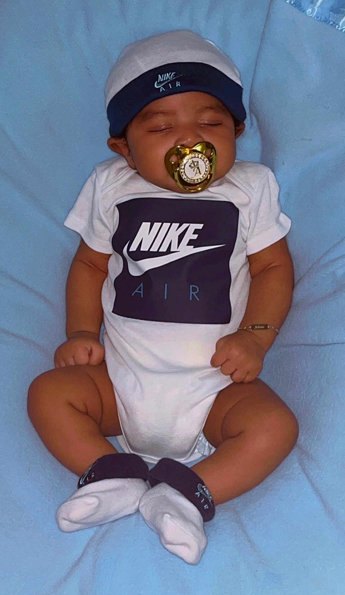 Adonis is registered to the contest to win money with this photo: baby, infant, sleeping, pacifier, hat, nike_air, onesie, socks, bracelet, blue_blanket, clothing, footwear, headwear, cute, child, resting, portrait, indoors, soft_texture, peaceful