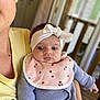baby, infant, headband, bib, polka_dots, knit_sweater, adult, holding, indoor, window, blurred_background, curious, portrait, child, person, closeup, soft_light, casual_clothing, yellow_shirt, face