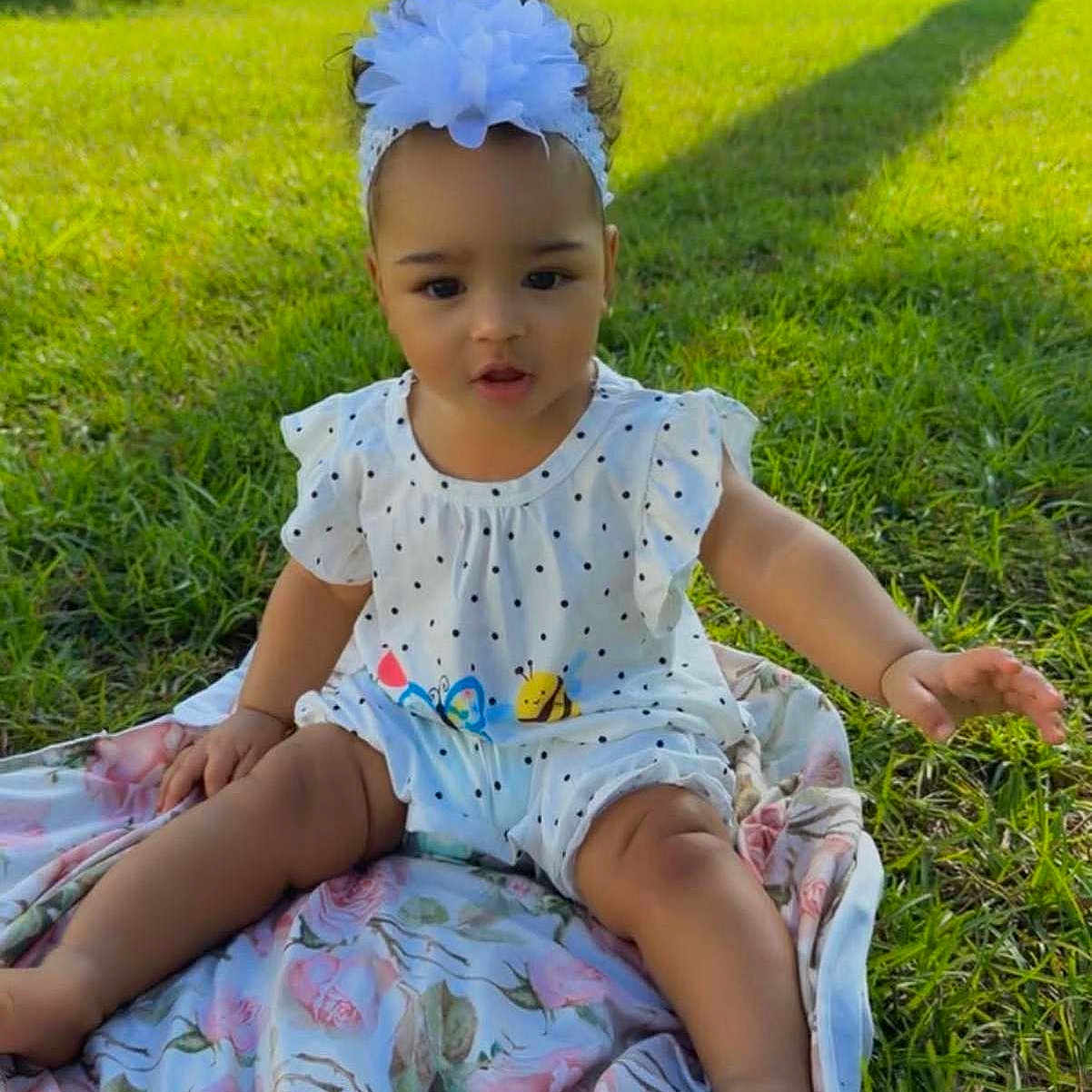 Raelynn joined the competition — help win amazing prizes! baby, child, toddler, sitting, outdoors, grass, garden, blanket, floral, dress, polka_dot, headband, blue_flower, portrait, face, person, sunlight, trees, backyard, cute