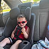 toddler, child, car_seat, sunglasses, red, seatbelt, car_interior, window, sunlight, smile, shorts, clothing, seat, safety, person, happy, casual, hair, skin, daylight