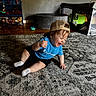 toddler, child, laughing, blue_shirt, baseball_cap, sitting, rug, indoor, toy_shelf, playpen, wooden_floor, smiling, person, happy, casual_clothing, sock, home, carpet, furniture, playroom