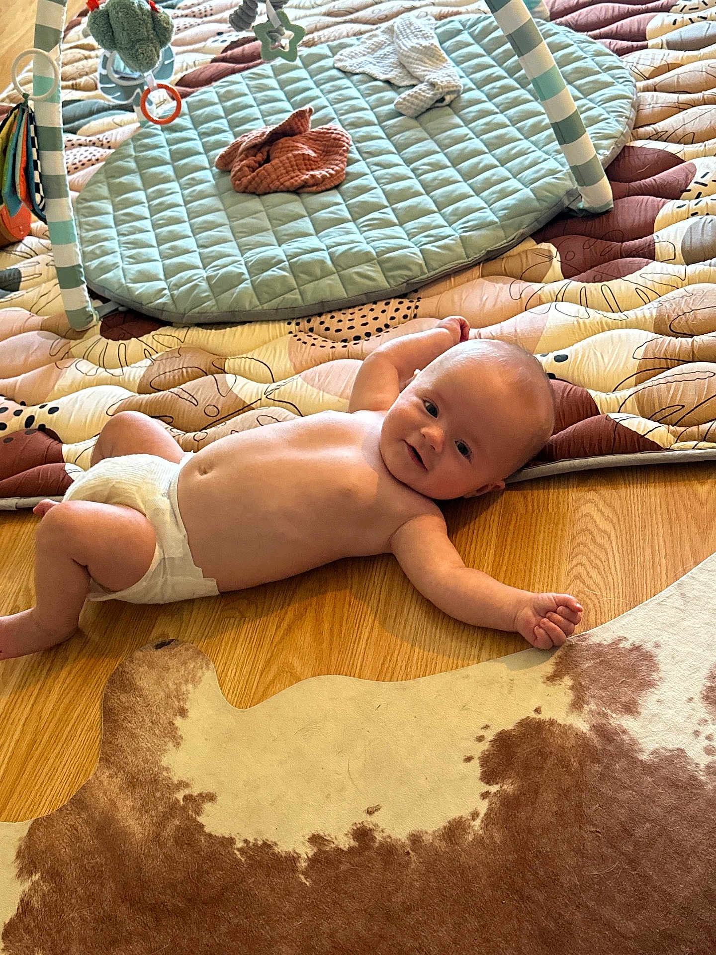 Blake is registered to the contest to win money with this photo: baby, diaper, floor, wooden_floor, rug, play_mat, toy, infant, child, lying_down, smiling, indoor, cute, patterned_rug, quilted_mat, curious, person, skin, head, arm