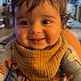 baby, child, smile, indoor, cozy, bib, clothing, curly_hair, face, happy, person, portrait, infant, cute, warm_lighting, home, expression, young, sitting, closeup