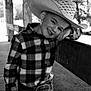 child, boy, cowboy_hat, plaid_shirt, belt_buckle, jeans, smile, portrait, outdoor, wooden_fence, black_and_white, happy, leaning, hat, young, person, casual, ranch, nature, sunlight