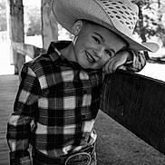 Landon is registered to the contest to win money with this photo: child, boy, cowboy_hat, plaid_shirt, belt_buckle, jeans, smile, portrait, outdoor, wooden_fence, black_and_white, happy, leaning, hat, young, person, casual, ranch, nature, sunlight