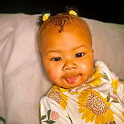 Essence is registered to the contest to win money with this photo: baby, child, tongue_out, hair_clips, curly_hair, yellow, sunflower_pattern, onesie, face, cute, infant, portrait, smiling, playful, indoors, closeup, skin, expression, lying_down, soft_surface