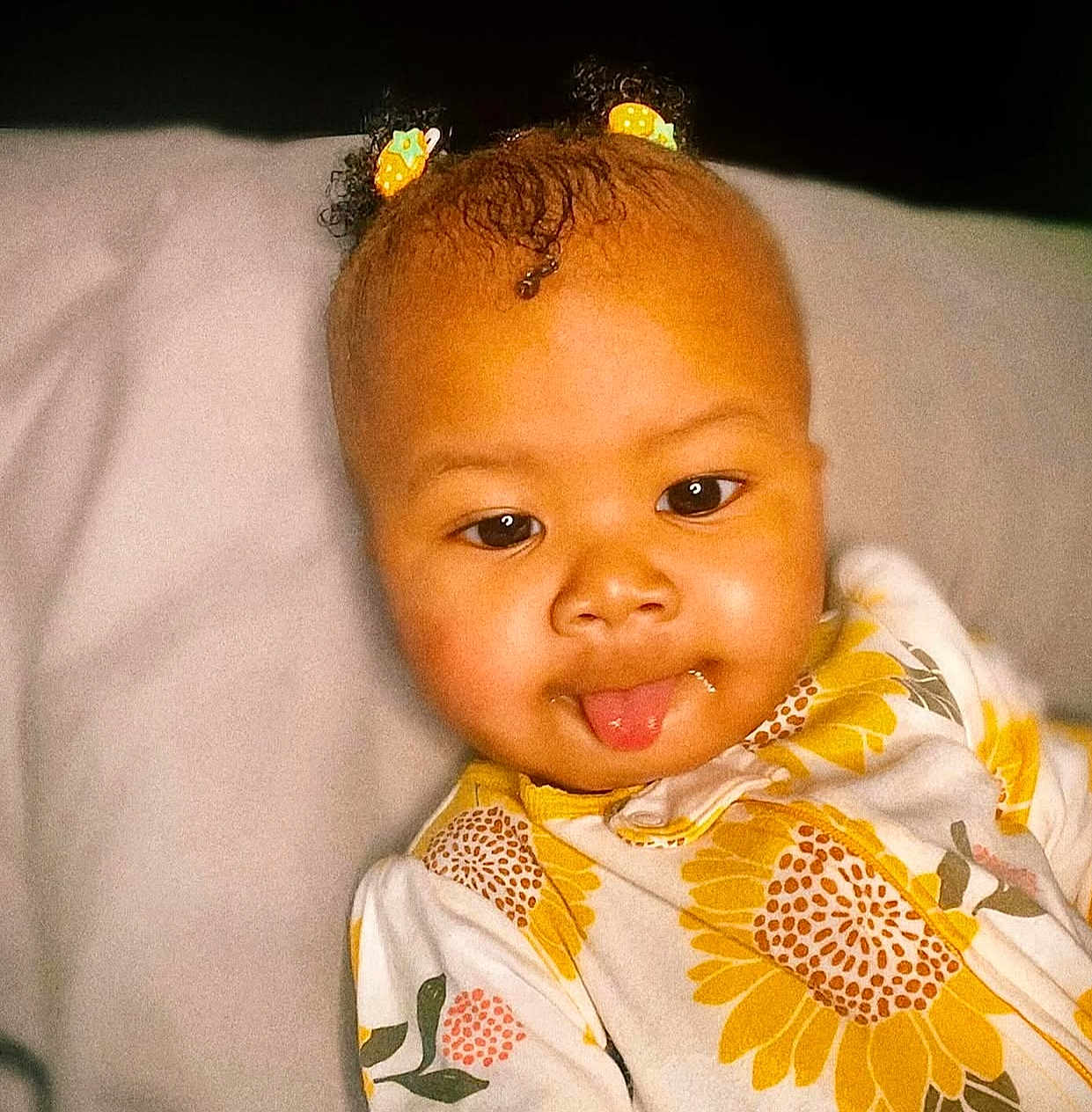 Essence is registered to the contest to win money with this photo: baby, child, tongue_out, hair_clips, curly_hair, yellow, sunflower_pattern, onesie, face, cute, infant, portrait, smiling, playful, indoors, closeup, skin, expression, lying_down, soft_surface